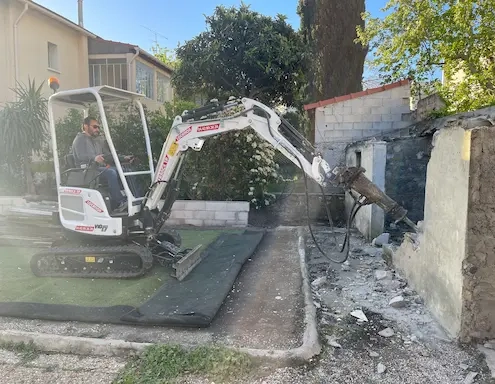 Mini-pelle démolissant un mur dans un jardin.