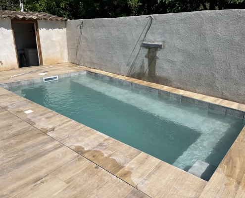 Piscine rectangulaire mise en eau, bordée d’une terrasse à l’aspect bois entre des murs maçonnés.