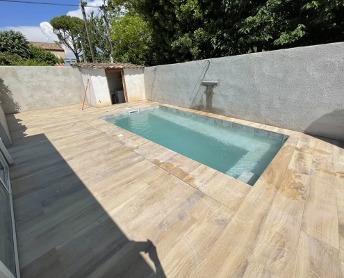 Terrasse à l’aspect bois entourant une piscine rectangulaire dans un jardin délimité par des murs.